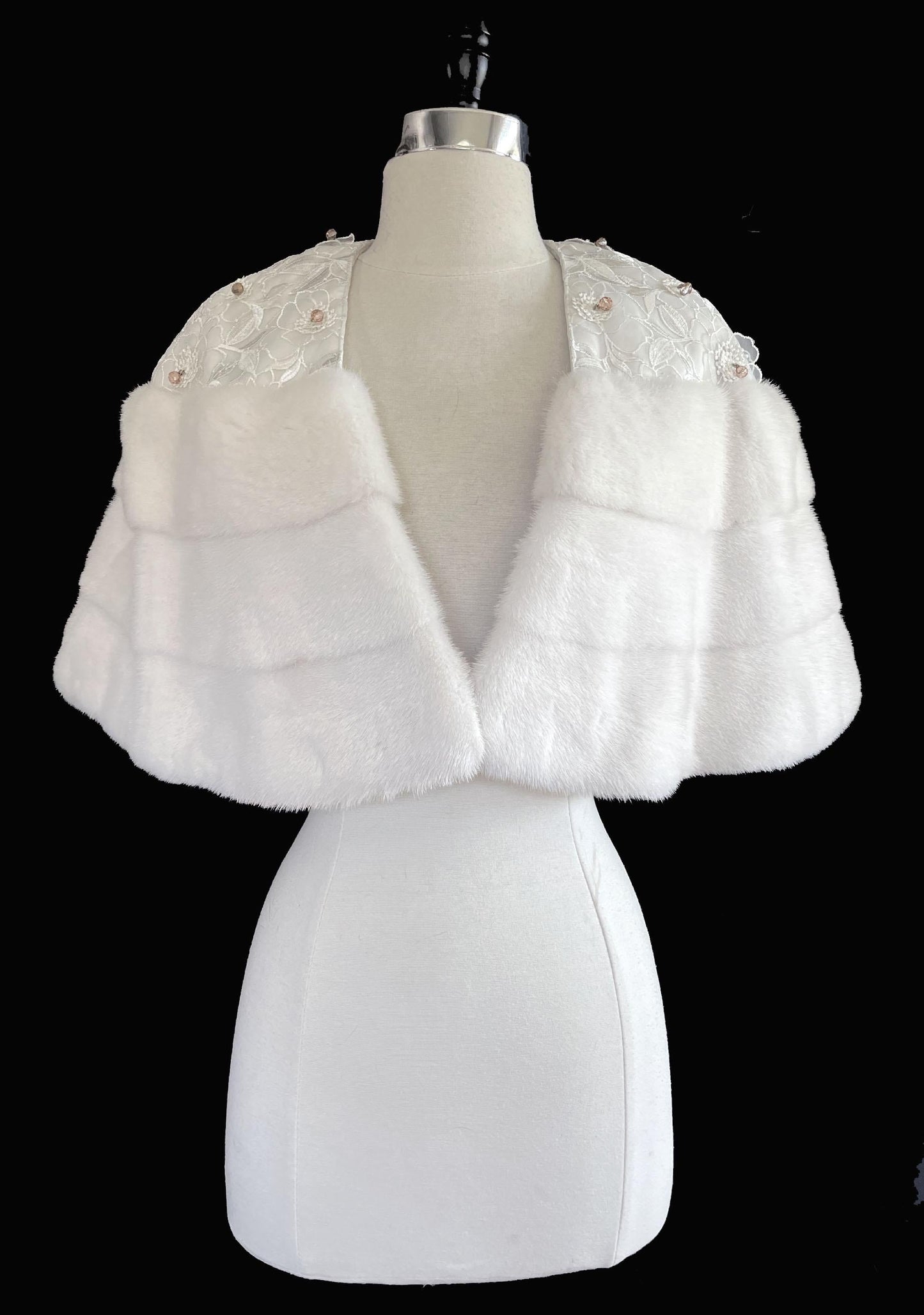 May include: A white mink cape displayed on a mannequin. The cape features a layered design with horizontal bands of fur. The shoulders are adorned with white lace and small, decorative embellishments. 