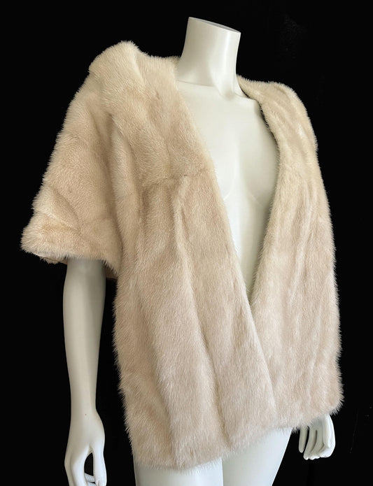 May include: A light beige fur cape with short sleeves. The cape is displayed on a mannequin against a black background. The text "find your dream fur at MOXIEFURS.COM" is at the bottom of the image.