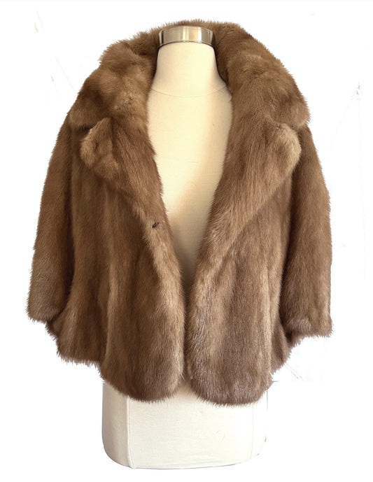 May include: A brown fur jacket with a wide collar and short sleeves is displayed on a mannequin. The jacket has a single button closure and a soft, textured appearance. The text "find your dream fur at MOXIEFURS.COM" is at the bottom.